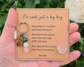May include: A keyring featuring a wooden charm engraved with a paw print and the name Titch, alongside three beads in green, white, and pink, attached to a card with the text No words, just a big hug and a small pink paw-shaped stone.