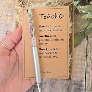 May include: A silver-toned ballpoint pen with a clear barrel filled with small purple, pink, and white crystals. The pen is clipped to a rectangular kraft paper card labelled Teacher, listing properties for Fluorite, Amethyst, and Rose Quartz.