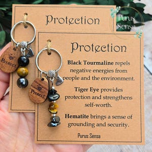 Op de afbeelding: Een bruine kaart met de tekst "Protection" en een beschrijving van de beschermende eigenschappen van zwarte toermalijn, tijgeroog en hematiet. De kaart wordt vergezeld door een sleutelhanger met een houten bedel, zwarte toermalijn, tijgeroog en hematiet kralen.