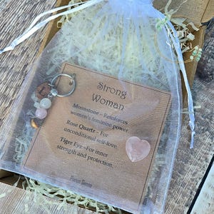 May include: A gift set in a white mesh bag, featuring a keychain with beads and a card that reads "Strong Woman." The card lists the properties of moonstone, rose quartz, and tiger eye. A heart-shaped rose quartz is also included.