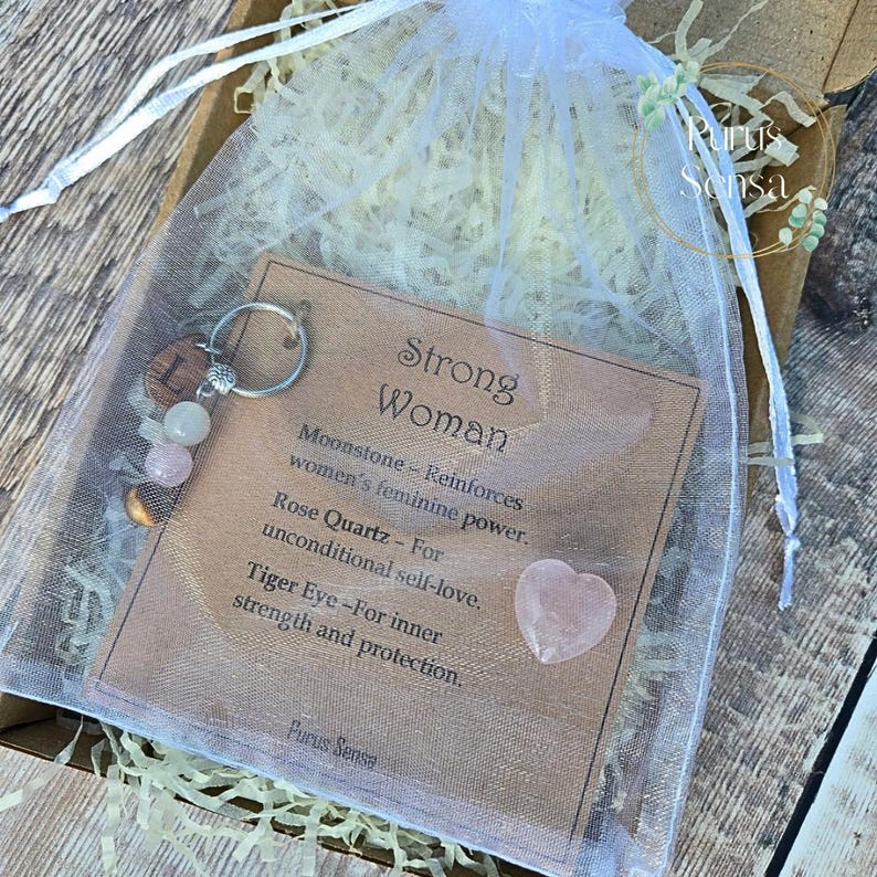 May include: A gift set in a white mesh bag, featuring a keychain with beads and a card that reads "Strong Woman." The card lists the properties of moonstone, rose quartz, and tiger eye. A heart-shaped rose quartz is also included.