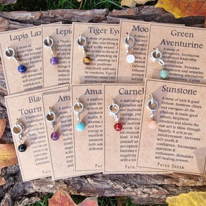 May include: A set of ten charm pendants with different colored gemstones and descriptions of their properties. The charms are made of silver and have a ring attached to the top. The descriptions are printed on brown cards with white text.