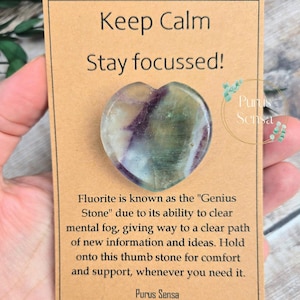 May include: A heart-shaped fluorite crystal thumb stone with green, purple, and white colours. The stone is on a brown card with the text "Keep Calm Stay focussed!" and "Fluorite is known as the "Genius Stone" due to its ability to clear mental fog, giving way to a clear path of new information and ideas. Hold onto this thumb stone for comfort and support, whenever you need it. Purus Sensa".