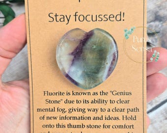 May include: A heart-shaped fluorite crystal thumb stone with green, purple, and white colors. The stone is on a brown card with the text "Keep Calm Stay focussed!" and "Fluorite is known as the "Genius Stone" due to its ability to clear mental fog, giving way to a clear path of new information and ideas. Hold onto this thumb stone for comfort and support, whenever you need it. Purus Sensa".
