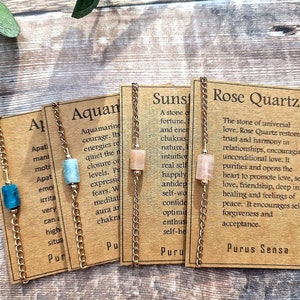 May include: Four gold chain bracelets with different colored gemstones and corresponding descriptions on cards. The cards describe the properties of the stones: Aquamarine, Sunstone, Rose Quartz, and Apatite.