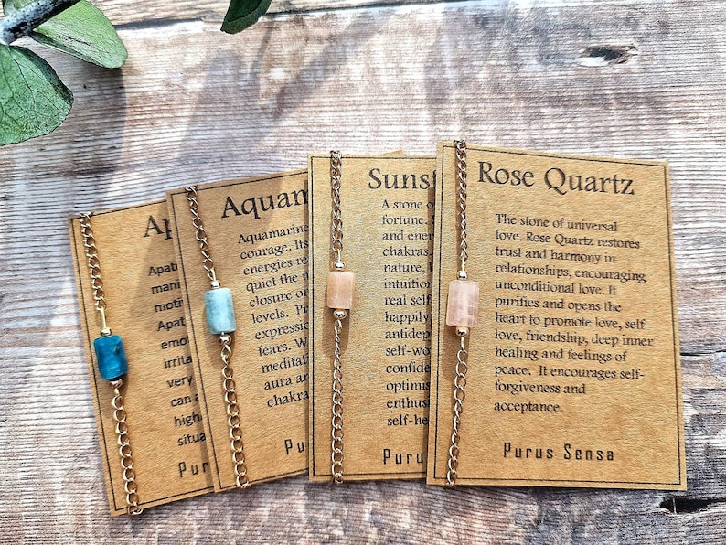 May include: Four gold chain bracelets with different colored gemstones and corresponding descriptions on cards. The cards describe the properties of the stones: Aquamarine, Sunstone, Rose Quartz, and Apatite.