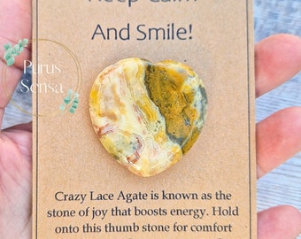 May include: A heart-shaped Crazy Lace Agate crystal with brown, yellow, and white patterns. The crystal is on a brown card with the text "Keep Calm And Smile!" and "Crazy Lace Agate is known as the stone of joy that boosts energy. Hold onto this thumb stone for comfort and support whenever you need it. Purus Sensa".