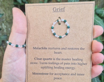 May include: A beaded ring with clear and green beads is displayed on a brown card. The card reads "Grief" and includes text about the healing properties of malachite, clear quartz, and moonstone. The brand name "Purus Sensa" is at the bottom.