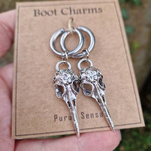 May include: A pair of silver hoop earrings with skull-shaped charms. The charms have a floral design and a pointed beak. The earrings are labeled "Boot Charms" and "Puru Sensa".