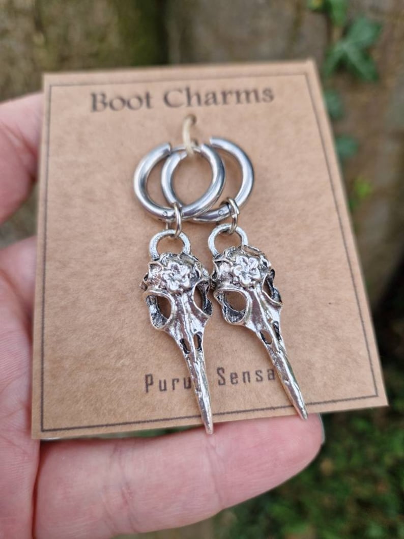 May include: A pair of silver hoop earrings with skull-shaped charms. The charms have a floral design and a pointed beak. The earrings are labeled "Boot Charms" and "Puru Sensa".