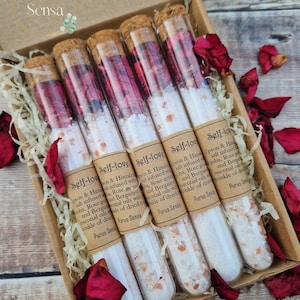 May include: Set of six glass test tubes filled with white bath salts and rose petals. Each tube has a cork stopper and a label that reads "Self-Love" with a description of the ingredients. The tubes are arranged in a box with shredded paper and rose petals.