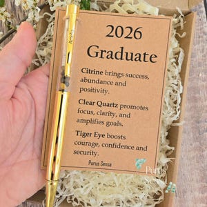 Puede incluir: Un bolígrafo dorado con un barril transparente lleno de pequeñas piedras, presentado en una caja de regalo con una tarjeta. La tarjeta dice "2026 Graduate" y enumera los beneficios de la citrina, el cuarzo transparente y el ojo de tigre. El bolígrafo está etiquetado "Amelia".