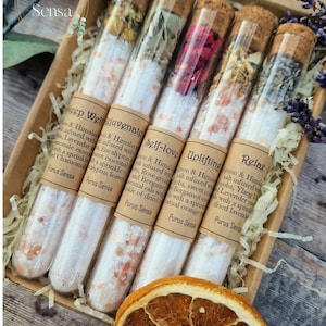 Könnte beinhalten: Satz mit sechs Glas-Reagenzgläsern, gefüllt mit Badesalz und getrockneten Blumen. Die Röhrchen sind mit den Worten "Deep Well Rejuvenate", "Self-love", "Uplifting" und "Relax" beschriftet. Die Etiketten enthalten auch den Markennamen "Purus Sensa".