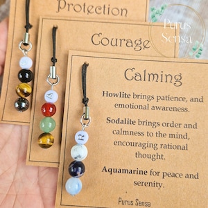 Op de afbeelding: Drie bruine kartonnen kaartjes met zwart touw en kristallen kralenbedels. De kaartjes zijn gelabeld met "Bescherming", "Moed" en "Kalmerend". Het kaartje "Kalmerend" heeft tekst die de eigenschappen van howliet, sodaliet en aquamarijn kristallen beschrijft.