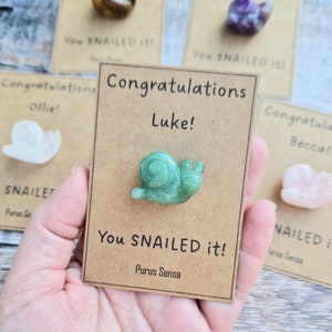 May include: A brown card with the text "Congratulations Luke! You SNAILED it! Purus Sensa" and a green snail figurine.