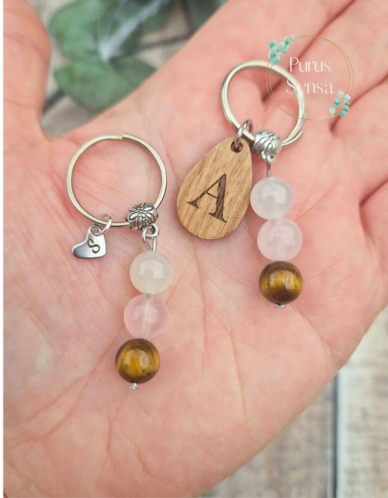 May include: Two keychains with silver rings. One keychain has a wooden teardrop with the letter "A" engraved. Both keychains have a floral charm and feature a series of beads in white, pink, and brown. One keychain has a heart charm with the letter "S".
