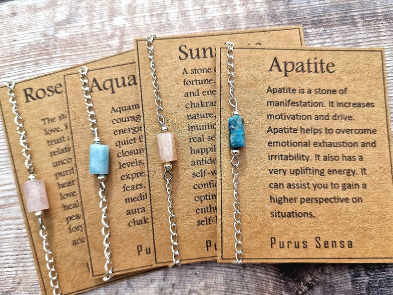 May include: Four silver chain bracelets with different colored gemstone beads and corresponding cards with the names "Rose", "Aqua", "Sun", and "Apatite". Each card describes the stone's properties and meaning.