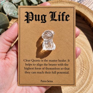 Könnte beinhalten: Eine kleine, klare Quarzschnitzerei in Form eines Mopses, befestigt auf einer rechteckigen, beigefarbenen Präsentationskarte. Die Karte zeigt oben den schwarzen Text "Pug Life" und darunter einen Absatz über klaren Quarz.