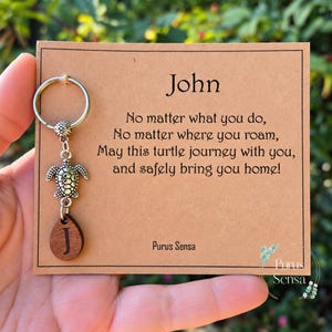 May include: A silver-toned metal keyring featuring a turtle charm and a small wooden teardrop-shaped pendant with the letter J. The keyring is attached to a tan card with the text John and a poem about a turtle journeying and returning home.