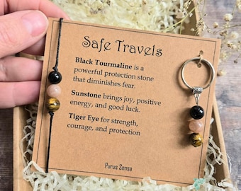 May include: A black cord bracelet and a silver-toned metal keyring, both featuring three beads of black tourmaline, sunstone, and tiger eye. They are attached to a tan card with the text Safe Travels and descriptions of the stones.