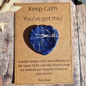 May include: A blue heart-shaped sodalite crystal with white veins. The crystal is on a brown card with the text "Keep Calm You've got this!" and "Sodalite brings order and calmness to the mind. Hold onto this thumb stone for comfort and support whenever you need it. Purus Sensa."