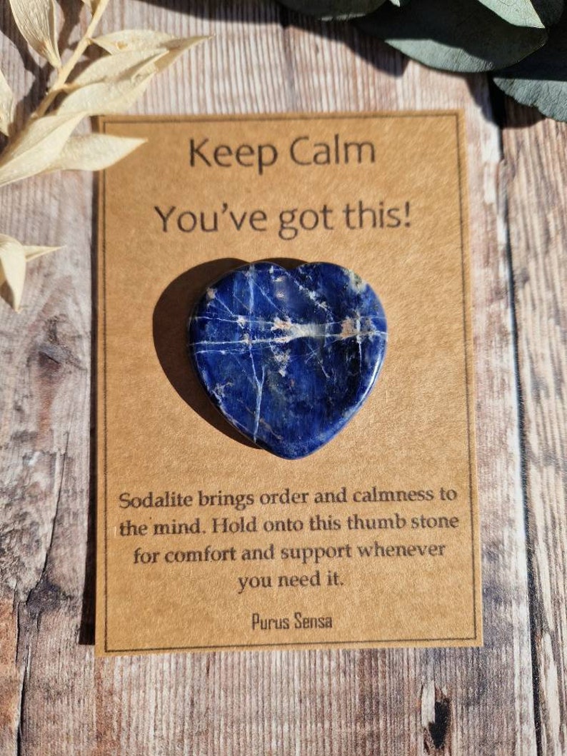 May include: A blue heart-shaped sodalite crystal with white veins. The crystal is on a brown card with the text "Keep Calm You've got this!" and "Sodalite brings order and calmness to the mind. Hold onto this thumb stone for comfort and support whenever you need it. Purus Sensa."