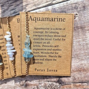May include: A gold chain necklace with aquamarine stones and a brown card with the text "Aquamarine is a stone of courage. Its calming energies reduce stress and quiet the mind. Useful for closure on all levels. Promotes self-expression and soothes fears. Wonderful for meditation. Shields the aura and aligns the chakras. Purus Sensa"