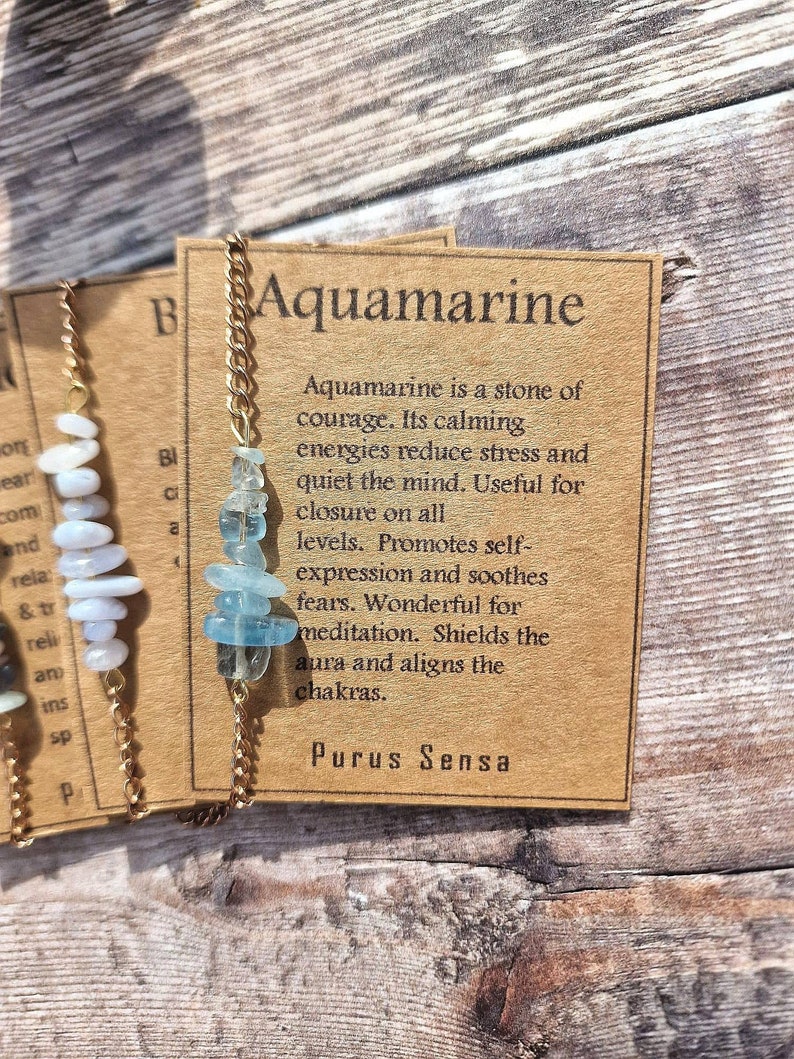 May include: A gold chain necklace with aquamarine stones and a brown card with the text "Aquamarine is a stone of courage. Its calming energies reduce stress and quiet the mind. Useful for closure on all levels. Promotes self-expression and soothes fears. Wonderful for meditation. Shields the aura and aligns the chakras. Purus Sensa"