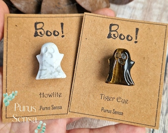 May include: Two small ghost-shaped figurines made of Howlite and Tiger Eye. The figurines are on brown cards with the word "Boo!" written on them. The cards also have the names of the stones and the brand "Purus Sensa" written on them.