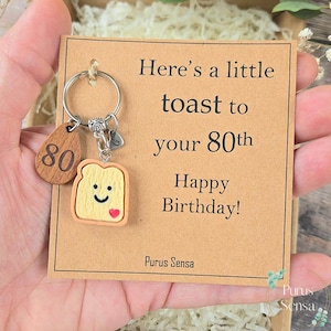 Puede incluir: Un llavero con un dije de madera con el número 80 y un dije en forma de tostada con una cara sonriente y un corazón. La tarjeta dice "Here's a little toast to your 80th Happy Birthday!"
