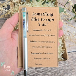Op de afbeelding: Zilveren pen met een transparante huls gevuld met blauwe en groene edelstenen. De pen ligt naast een kaart met de tekst "Something blue to sign 'I do'" en beschrijvingen van edelstenen. Een bruiloftaccessoire.