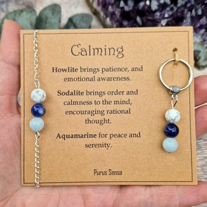 May include: A calming crystal necklace with howlite, sodalite, and aquamarine beads. The necklace is on a brown card with the text "Calming" and descriptions of the crystals' properties. The text also includes the phrase "Purus Sensa".