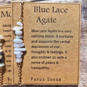 May include: A gold chain necklace with a blue lace agate pendant. The pendant is made of several small, flat, light blue stones. The card attached to the necklace reads "Blue Lace Agate is a very calming stone. It nurtures and supports the verbal expression of our thoughts & feelings. It also endows us with a sense of peace & tranquility. Purus Sensa."