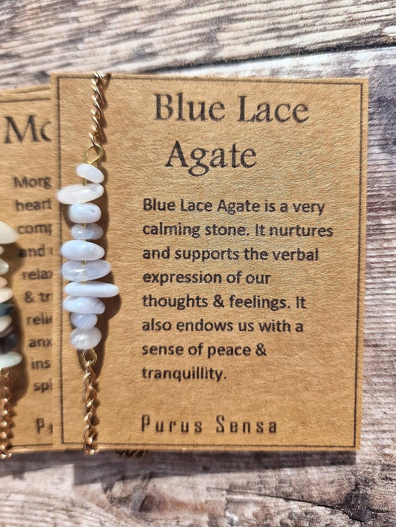 May include: A gold chain necklace with a blue lace agate pendant. The pendant is made of several small, flat, light blue stones. The card attached to the necklace reads "Blue Lace Agate is a very calming stone. It nurtures and supports the verbal expression of our thoughts & feelings. It also endows us with a sense of peace & tranquility. Purus Sensa."
