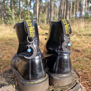Könnte beinhalten: Schwarze Lederstiefel mit gelben Schnürsenkeln und silbernen Halbmond-Anhängern. Die Anhänger haben einen blauen Edelstein in der Mitte.
