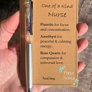 Op de afbeelding: Zilveren pen met een doorzichtige cilinder gevuld met amethist, fluoriet en rozenkwarts kristallen. De pen heeft een kaart eraan bevestigd met de tekst "One of a Kind Nurse" en een lijst met de kristallen en hun eigenschappen. De kaart heeft ook de tekst "Purus Sensa".