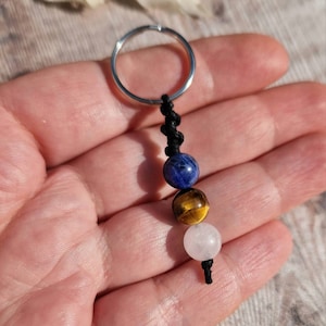 May include: A keychain with a silver ring and a black cord holding three gemstone beads. The beads are blue, gold, and white. The keychain is held in a hand.