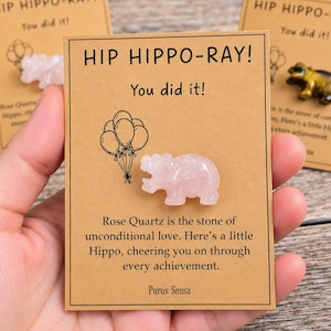 May include: A light pink rose quartz hippo figurine attached to a rectangular tan card. The card features the text HIP HIPPO-RAY! You did it! and a line drawing of balloons. Below the hippo, text reads Rose Quartz is the stone of unconditional love.