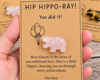 May include: A light pink rose quartz hippo figurine attached to a rectangular tan card. The card features the text HIP HIPPO-RAY! You did it! and a line drawing of balloons. Below the hippo, text reads Rose Quartz is the stone of unconditional love.