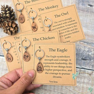 Könnte beinhalten: Holz-Schlüsselanhänger mit silbernen Ringen, jeweils mit einer Gravur eines anderen Tieres: Schmetterling, Affe, Eule, Huhn, Adler und Hirsch. Die Schlüsselanhänger werden mit Informationskarten präsentiert.