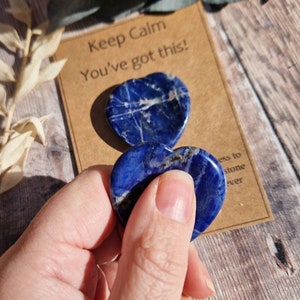 May include: Two blue heart-shaped lapis lazuli stones with white veins. The stones are held in a hand. A card with the text "Keep Calm You've got this!" is in the background.