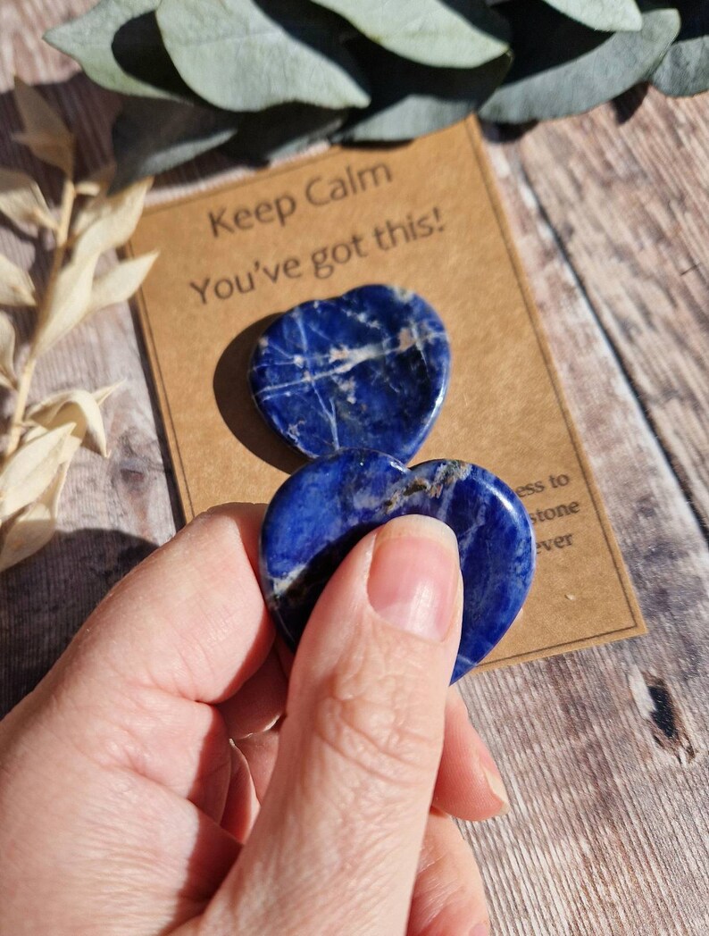 May include: Two blue heart-shaped lapis lazuli stones with white veins. The stones are held in a hand. A card with the text "Keep Calm You've got this!" is in the background.