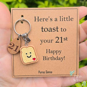 May include: A keyring with a wooden tag showing the number 21, and a toast-shaped charm with a smiling face and a red heart. The card reads "Here's a little toast to your 21st Happy Birthday!" and has the brand name "Purus Sensa".