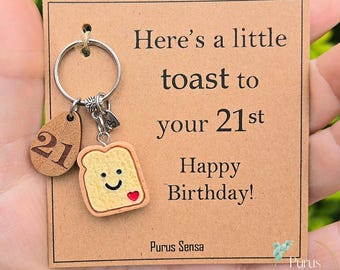 Könnte beinhalten: Ein Schlüsselanhänger mit einem Holzanhänger mit der Zahl 21 und einem toastförmigen Anhänger mit einem lächelnden Gesicht und einem roten Herz. Die Karte lautet "Here's a little toast to your 21st Happy Birthday!" und hat den Markennamen "Purus Sensa".