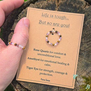 Könnte beinhalten: Ein Ring aus Rosenquarz-, Amethyst- und Tigeraugenperlen. Der Ring wird mit einer Karte gezeigt, auf der steht: "Life is tough... But so are you!" und die Eigenschaften der Steine beschreibt.