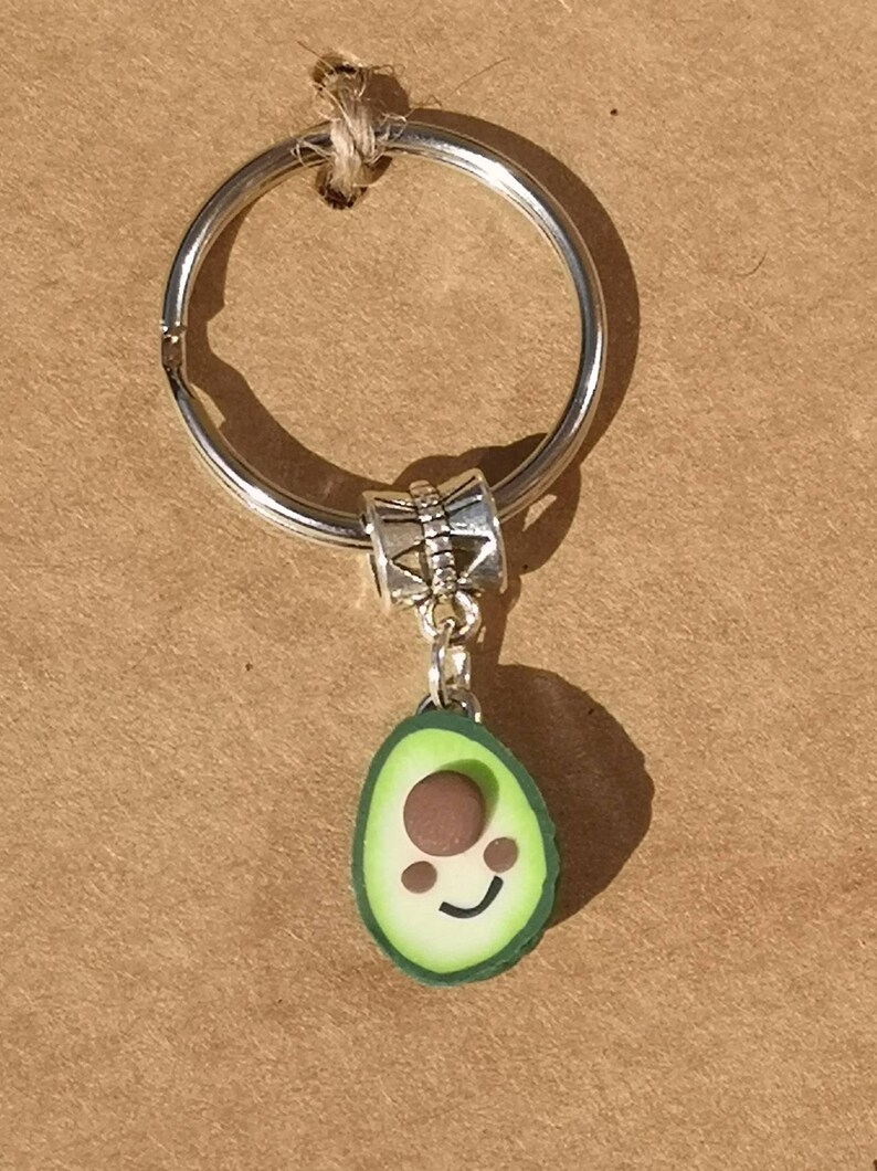 May include: A silver key chain with a green avocado charm. The avocado has a brown pit and a smiling face.