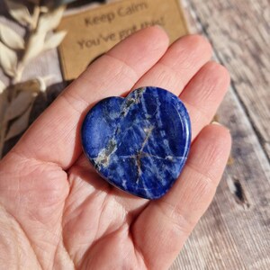 May include: A heart-shaped blue sodalite crystal with white and gold veins. The crystal is being held in a hand.