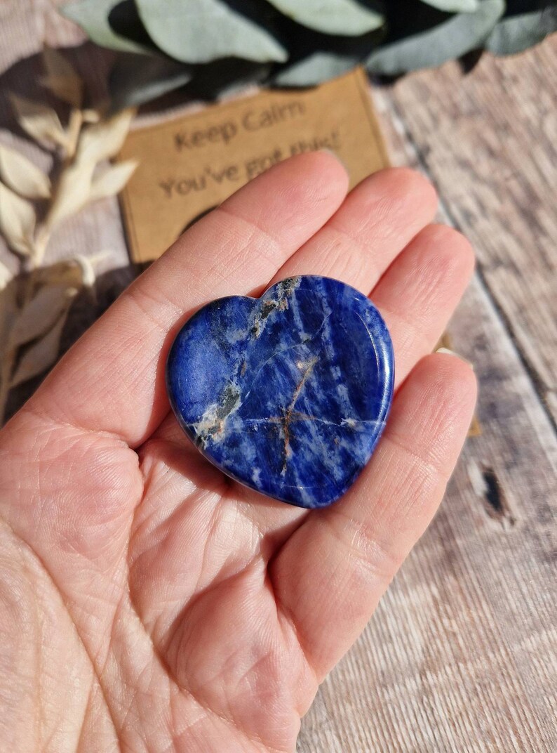 May include: A heart-shaped blue sodalite crystal with white and gold veins. The crystal is being held in a hand.