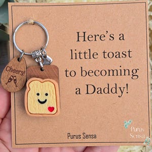 May include: A metal keychain featuring a wooden charm engraved with Cheers! and two champagne glasses, alongside a toast-shaped charm with a smiling face and a red heart. The keychain is attached to a card with the text Here's a little toast to becoming a Daddy! and Purus Sensa.