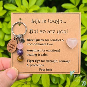 Peut inclure: Un porte-clés avec une étiquette en bois avec l'inscription "You've got this" et un cœur, ainsi que trois perles de pierres précieuses. La carte indique "Life is tough... But so are you!" et énumère les propriétés du quartz rose, de l'améthyste et de l'œil de tigre.